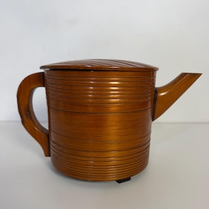 May include: A wooden teapot with a lid, handle, and spout. The teapot is a warm brown color and has horizontal grooves around the body. The lid has a ridged design. The handle is curved, and the spout extends from the side.