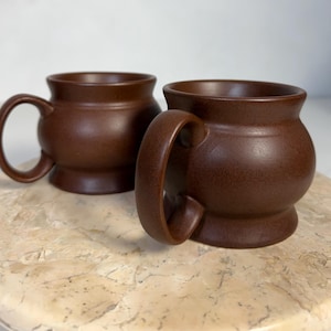 Puede incluir: Dos tazas de cerámica marrón con cuerpos redondeados y asas grandes. Las tazas tienen una textura moteada y se asientan sobre una superficie clara y texturizada. Las tazas son de forma y tamaño similares.