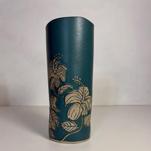 Könnte beinhalten: Türkisfarbene Keramikvase mit geprägtem Hibiskusblüten-Design. Die Vase ist zylindrisch mit leicht gebogenem Oberteil. Die Blüten sind hellbeige und die Blätter detailliert.