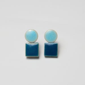 May include: A pair of earrings with a light blue circular top and a dark blue square base. The earrings have a glossy finish and are set against a white background. The design is minimalist and geometric.
