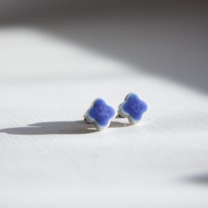 Könnte beinhalten: Ein Paar kleiner Ohrstecker aus Keramik. Jeder Ohrring hat ein vierblättriges Kleeblatt-Design mit einem leuchtend blauen Zentrum und einem weißen Rand. Die Ohrringe sind vor einem weißen Hintergrund mit Schatten platziert.
