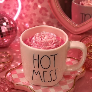 Op de afbeelding: Een witte keramische mok met de zwarte tekst "HOT MESS", met een roze roosvormige kaars. De mok staat op een roze en wit geblokt dienblad met een gouden rand. Een roze discobal en een hartvormige spiegel staan op de achtergrond.