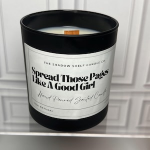 Peut inclure: Une bougie noire avec une étiquette blanche portant l'inscription "Spread Those Pages Like A Good Girl". La bougie est coulée à la main et parfumée, avec une mèche en bois. L'étiquette indique également "The Shadow Shelf Candle Co." et "100% Natural."