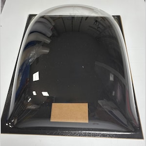 May include: A clear, domed plastic cover with a black base. The cover is rectangular with rounded edges and a small, rectangular cardboard piece adhered to the base. The cover is reflective, showing distorted reflections.