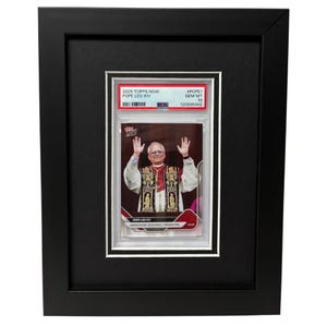 May include: Framed 2025 Topps Now Pope Leo XIV trading card. The card is encased in a protective holder and features a photo of the Pope with his hands raised. The frame is black with a white mount.