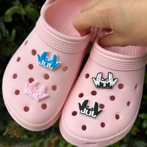 Peut inclure: Deux sabots roses ornés de breloques. L'une des chaussures présente une breloque bleue et blanche, l'autre une breloque noire et blanche. Les breloques représentent un geste de la main avec les lettres "JUL".