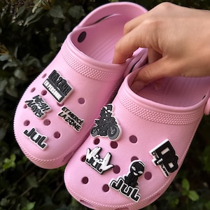 Peut inclure: Une paire de sabots rose clair décorés de divers charms noirs et blancs. Les charms portent les inscriptions "LA ZONE EN PERSONNE", "WEAH ALORS", "ET MERCÉ LA ZONE", "JUL" et "D'OR ET DE PLATINE", ainsi que des motifs de moto et de personnage.