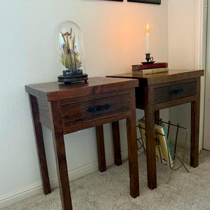 May include: Two dark brown wooden nightstands with drawers and black handles. One nightstand has a glass cloche with dried plants on top. The other has a lit candle and books. The nightstands are on a light carpet.