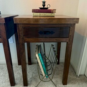 K&ouml;nnte beinhalten: Ein Paar rustikale Nachttische aus Holz mit Schublade und Zeitschriftenst&auml;nder. Jeder Tisch hat eine dunkelbraune Oberfl&auml;che, eine Schublade mit schwarzem Griff und einen Metall-Zeitschriftenst&auml;nder. Auf einem Tisch liegen B&uuml;cher und ein Kerzenhalter.