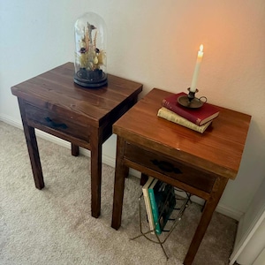 K&ouml;nnte beinhalten: Zwei Nachttische aus Holz mit Schubladen. Einer hat eine Glasglocke mit Trockenblumen, der andere B&uuml;cher und eine brennende Kerze. Die Tische sind dunkelbraun mit schwarzen Schubladengriffen und stehen auf einem hellen Teppich.