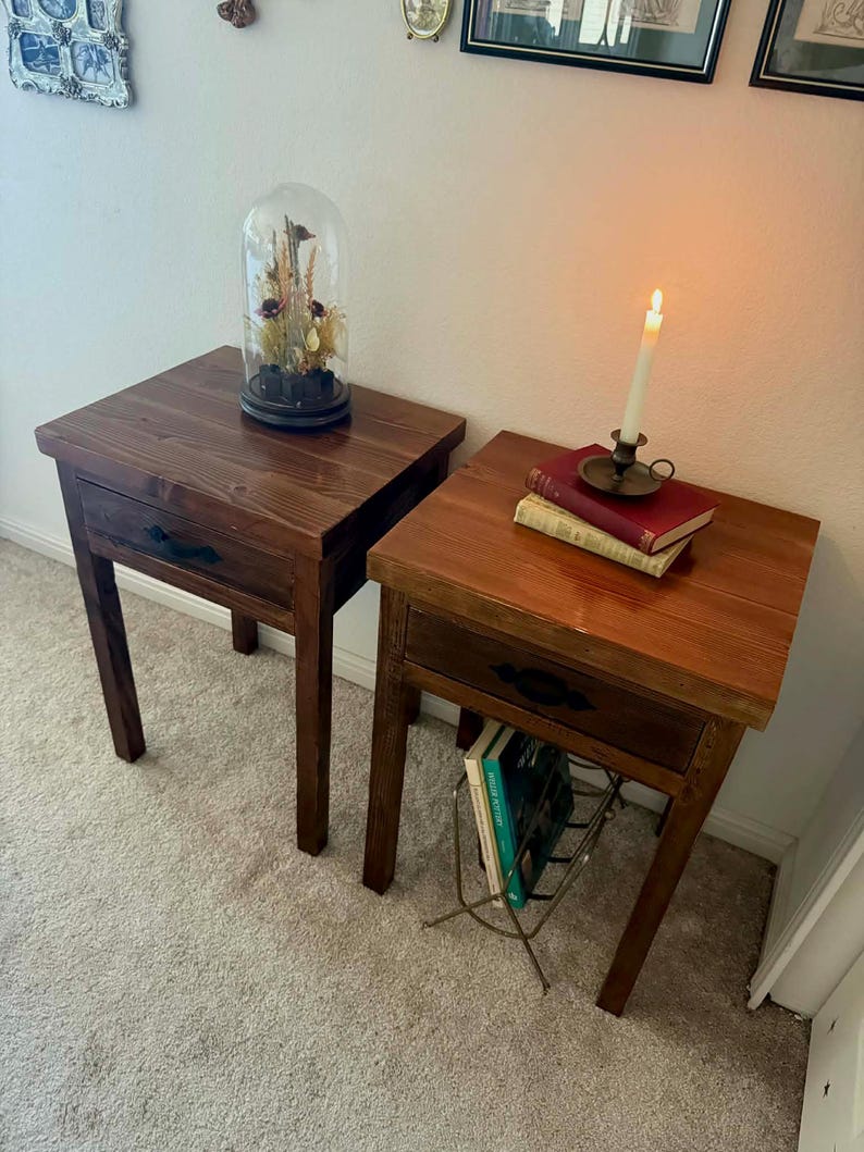 K&ouml;nnte beinhalten: Zwei Nachttische aus Holz mit Schubladen. Einer hat eine Glasglocke mit Trockenblumen, der andere B&uuml;cher und eine brennende Kerze. Die Tische sind dunkelbraun mit schwarzen Schubladengriffen und stehen auf einem hellen Teppich.