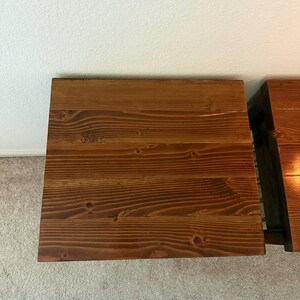 K&ouml;nnte beinhalten: Zwei Nachttische aus Holz mit dunkler brauner Beize. Die Tische sind rechteckig mit sichtbarer Holzmaserung und Knoten, was auf einen rustikalen Stil hindeutet. Die Tische stehen auf einem hellen Teppich.