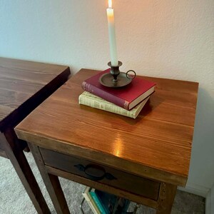 K&ouml;nnte beinhalten: Eine brennende wei&szlig;e Kerze in einem Messing-Kerzenhalter steht auf B&uuml;chern auf einem h&ouml;lzernen Beistelltisch. Der Tisch hat eine Schublade mit einem schwarzen Griff. Ein weiterer Holztisch ist links zu sehen.