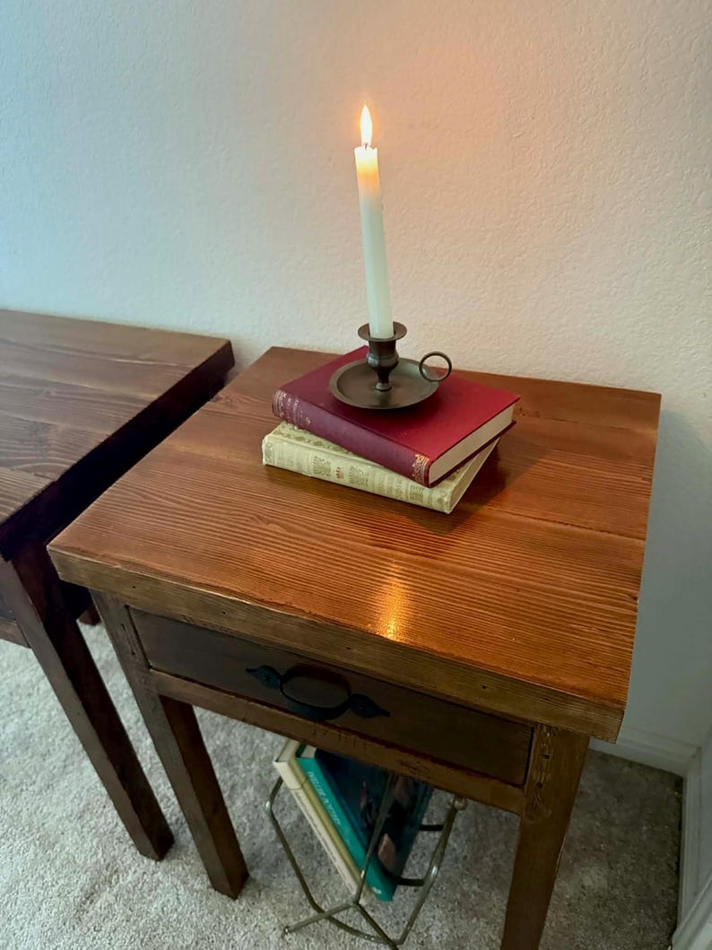 K&ouml;nnte beinhalten: Eine brennende wei&szlig;e Kerze in einem Messing-Kerzenhalter steht auf B&uuml;chern auf einem h&ouml;lzernen Beistelltisch. Der Tisch hat eine Schublade mit einem schwarzen Griff. Ein weiterer Holztisch ist links zu sehen.