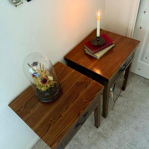 K&ouml;nnte beinhalten: Zwei Nachttische aus Holz mit warmem, r&ouml;tlich-braunem Finish. Ein Tisch enth&auml;lt eine Glasglocke mit getrockneten Blumen, der andere B&uuml;cher und eine brennende Kerze in einem Messinghalter. Die Tische haben dunkle Beschl&auml;ge.