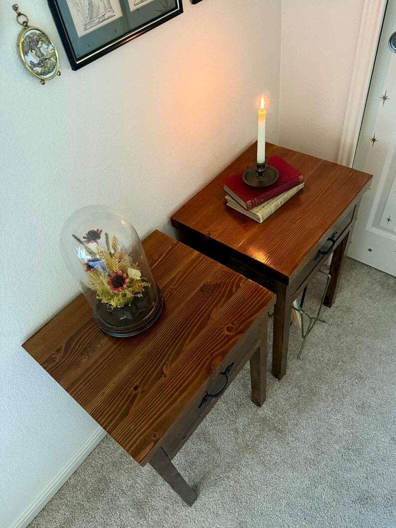K&ouml;nnte beinhalten: Zwei Nachttische aus Holz mit warmem, r&ouml;tlich-braunem Finish. Ein Tisch enth&auml;lt eine Glasglocke mit getrockneten Blumen, der andere B&uuml;cher und eine brennende Kerze in einem Messinghalter. Die Tische haben dunkle Beschl&auml;ge.
