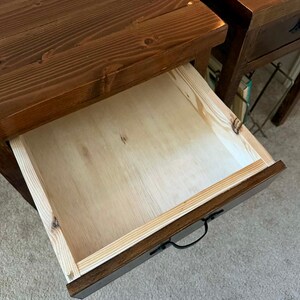 K&ouml;nnte beinhalten: Nahaufnahme eines Nachttisches aus Holz mit ge&ouml;ffneter Schublade. Der Nachttisch ist dunkelbraun gebeizt, mit sichtbarer Holzmaserung. Die Innenseite der Schublade ist helles Holz. Ein schwarzer Griff ist an der Schublade befestigt.
