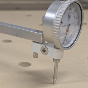 May include: A silver dial indicator with a white face and black markings, measuring up to 1.0 inches. The tool has a long, silver arm and a pointed tip, designed for precise measurements. The dial reads in increments of 0.001 inches.