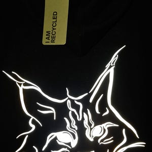 Pode incluir: Uma camisola preta com capuz apresentando um gráfico de linhas brancas de um rosto de gato com orelhas pontiagudas. Uma etiqueta retangular bege presa com um cordão diz I AM RECYCLED e écologie. O texto LINKON está impresso em branco abaixo do gato.
