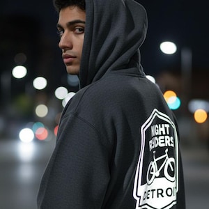 Puede incluir: Sudadera con capucha gris oscuro con un gráfico blanco en la espalda que dice "NIGHT RIDERS DETROIT" con una ilustración de bicicleta. La sudadera tiene una capucha con cordón. La persona está en una calle por la noche.