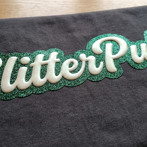 May include: A dark gray t-shirt with the words "Glitter Puff" in a white, cursive font. The text is outlined with green glitter and is centered on the shirt. The shirt is made of a soft material.