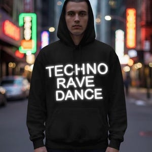 May include: Black hooded sweatshirt with the words "TECHNO RAVE DANCE" in white, block letters. The person is standing on a city street with neon signs in the background. The sweatshirt has a front pocket and a hood.