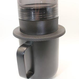 Puede incluir: Una cafetera negra con una parte superior cilíndrica transparente y un asa negra. La base tiene un diseño estriado, y el asa está unida a una estructura negra en forma de taza. La cafetera está diseñada para preparar café.