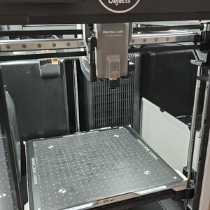 May include: A 3D printer with a black and silver frame. The printer features a build plate with a grid pattern and the text "320-325 mm". The printer has a display screen and the logo "Objects".