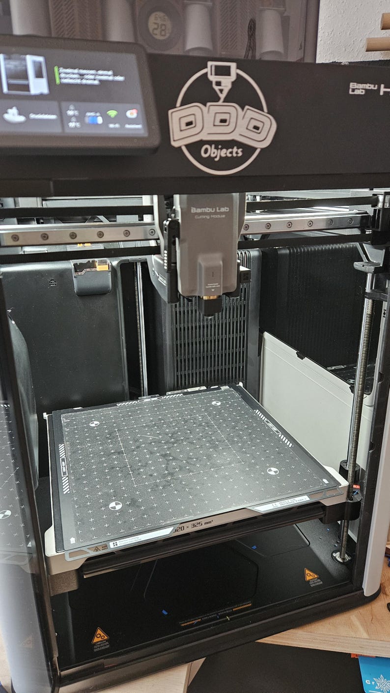 May include: A 3D printer with a black and silver frame. The printer features a build plate with a grid pattern and the text "320-325 mm". The printer has a display screen and the logo "Objects".
