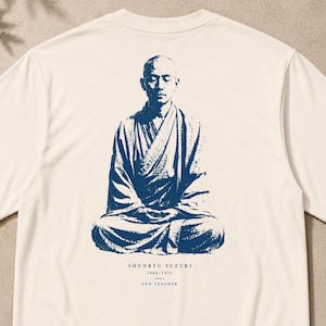 Peut inclure: Un t-shirt blanc cassé avec un motif bleu foncé représentant une personne en méditation vêtue de robes. Sous la figure, le texte indique : SHUNRYU SUZUKI, 1904-1971, ZEN TEACHER. Le motif est centré au dos du vêtement avec une impression détaillée.