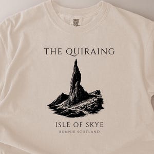 Pode incluir: Camiseta creme com uma ilustra&ccedil;&atilde;o em preto e branco de uma cordilheira. O texto "THE QUIRAING" est&aacute; acima da ilustra&ccedil;&atilde;o, e "ISLE OF SKYE BONNIE SCOTLAND" est&aacute; abaixo.