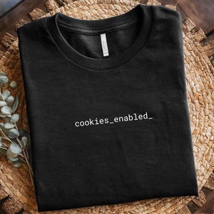 Könnte beinhalten: Schwarzes T-Shirt mit Rundhalsausschnitt und dem weißen Text "cookies_enabled_". Das Shirt besteht aus einem weichen Material und wird auf einer gewebten Oberfläche präsentiert. Ein Etikett ist am Kragen sichtbar.