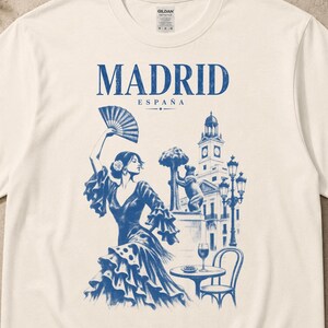 May include: A cream-coloured t-shirt featuring a blue graphic print. The text reads MADRID ESPAÑA. The illustration depicts a person dancing with a fan, the Bear and the Strawberry Tree statue, a street lamp, and a small table with a wine glass.