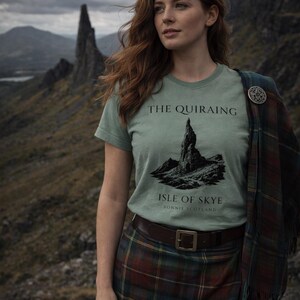 Pode incluir: Camiseta verde claro com o texto "THE QUIRAING ISLE OF SKYE BONNIE SCOTLAND" e um gr&aacute;fico de montanha. A pessoa est&aacute; usando uma saia xadrez e um xale xadrez com um broche de prata. O fundo apresenta uma paisagem montanhosa.