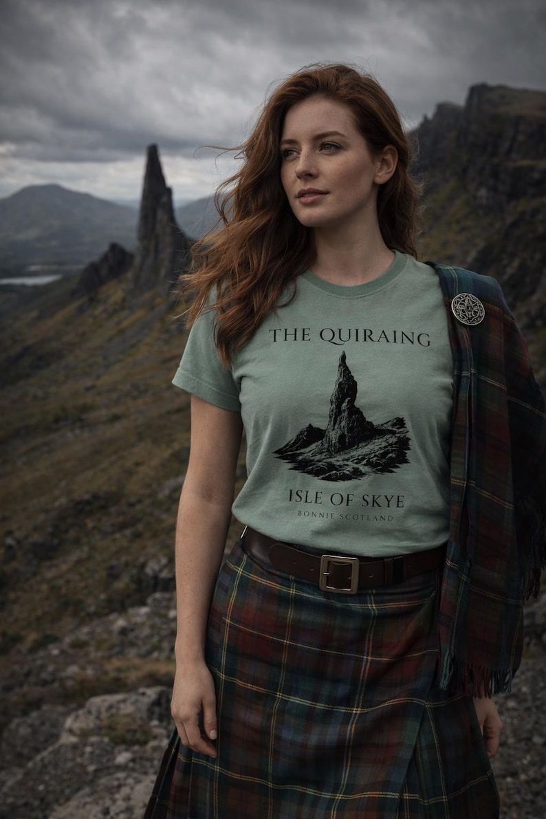 Pode incluir: Camiseta verde claro com o texto "THE QUIRAING ISLE OF SKYE BONNIE SCOTLAND" e um gr&aacute;fico de montanha. A pessoa est&aacute; usando uma saia xadrez e um xale xadrez com um broche de prata. O fundo apresenta uma paisagem montanhosa.