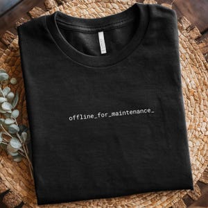 Può includere: Una maglietta nera con la scritta "offline_for_maintenance_" in bianco, centrata sul davanti. La maglietta è ben piegata e appoggiata su una superficie marrone intrecciata. La maglietta è realizzata in un materiale morbido.