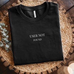 May include: Black t-shirt with the white text "USER NOT FOUND" across the front. The folded shirt rests on a circular woven mat, light brown in colour. Some green sprigs are visible to the left. The shirt is made of a soft material.