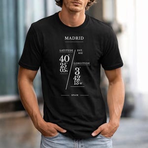 以下が含まれることがあります： 黒いTシャツに、白で「MADRID」の都市名がプリントされています。その下に「緯度」と「経度」の文字と座標があります。「SPAIN」の文字が下部にプリントされています。Tシャツは人が着ています。
