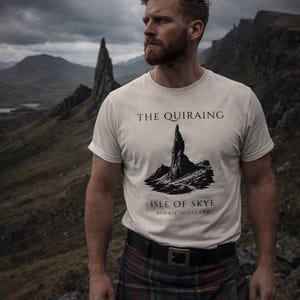 Pode incluir: Uma pessoa vestindo uma camiseta branca com um gr&aacute;fico de The Quiraing na Ilha de Skye. A pessoa tamb&eacute;m est&aacute; usando um kilt escoc&ecirc;s tradicional e um cinto preto. O fundo apresenta uma paisagem montanhosa sob um c&eacute;u nublado.