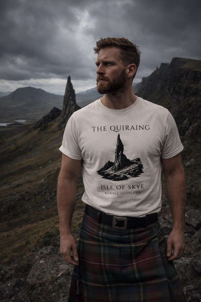 Pode incluir: Uma pessoa vestindo uma camiseta branca com um gr&aacute;fico de The Quiraing na Ilha de Skye. A pessoa tamb&eacute;m est&aacute; usando um kilt escoc&ecirc;s tradicional e um cinto preto. O fundo apresenta uma paisagem montanhosa sob um c&eacute;u nublado.