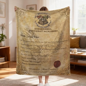 May include: A beige fleece blanket featuring a printed Hogwarts acceptance letter design. The graphic includes the Hogwarts crest, a large owl illustration, and text reading Hogwarts School of Witchcraft and Wizardry with a red wax seal.