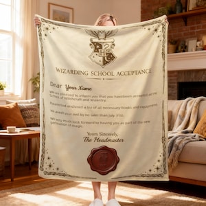 Puede incluir: Una manta de forro polar color crema con el estampado de una carta de aceptación a una escuela de magia. El diseño incluye un escudo con un búho, el texto "WIZARDING SCHOOL ACCEPTANCE", un espacio para un nombre y un sello de cera rojo.