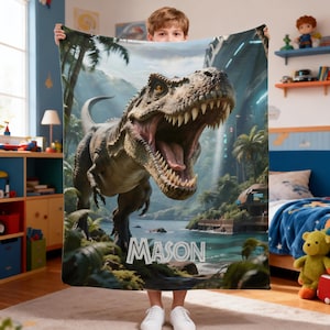 May include: A personalized blanket featuring a detailed illustration of a Tyrannosaurus Rex with an open mouth, set against a tropical landscape. The name "MASON" is printed in white. The blanket is held up by a person.