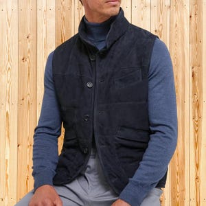 Puede incluir: Un chaleco de ante azul marino con diseño acolchado, cuello alzado y cierre de botones. El chaleco se lleva sobre un jersey de cuello alto azul. El fondo es una pared de madera.