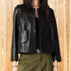 May include: A black leather jacket featuring a round neckline, long sleeves, and a front button closure. The jacket includes a single patch pocket on the right side with a button detail. It is worn over a black top and olive green trousers.