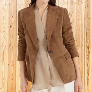 May include: A brown suede blazer featuring a single-button closure, notched lapels, and two front flap pockets. The blazer is worn over a light beige wrap-style top with long sleeves and a structured fit, showcasing a clean and classic design.