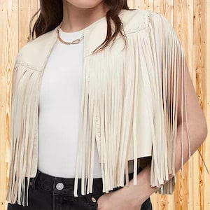 May include: A cream-colored cropped vest featuring long, flowing fringe detailing along the front panels and shoulders. The garment has a collarless, open-front design with decorative stitching along the edges, worn over a plain white top.