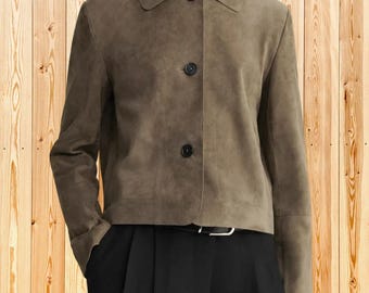 Op de afbeelding: Een olijfgroen suède jasje met een klassieke kraag, lange mouwen en een knoopsluiting aan de voorzijde met drie donkere knopen. Het jasje heeft een rechte zoom en wordt gedragen over een zwarte broek met een zichtbare zilveren riemgesp.