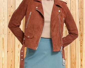 Op de afbeelding: Een roestkleurig suède bikerjack met een asymmetrische zilverkleurige ritssluiting aan de voorzijde, een reverskraag, een klein klepzakje en een riem met gesp aan de zoom. Het jack wordt gedragen over een beige top met col en een blauwe rok.