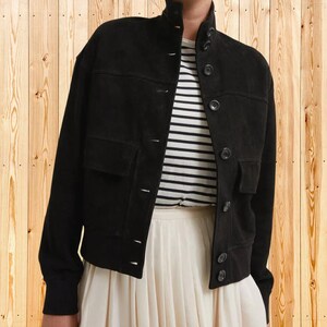 May include: A black suede jacket featuring a high collar, long sleeves, and a front button closure. The jacket includes two chest pockets with flaps and is worn over a white and black horizontal striped shirt and a white pleated skirt.